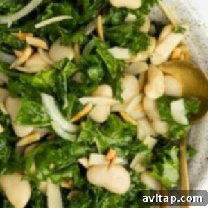A bowl of kale salad with white beans, sliced onions, slivered almonds, and shaved cheese, served with a gold fork and spoon.