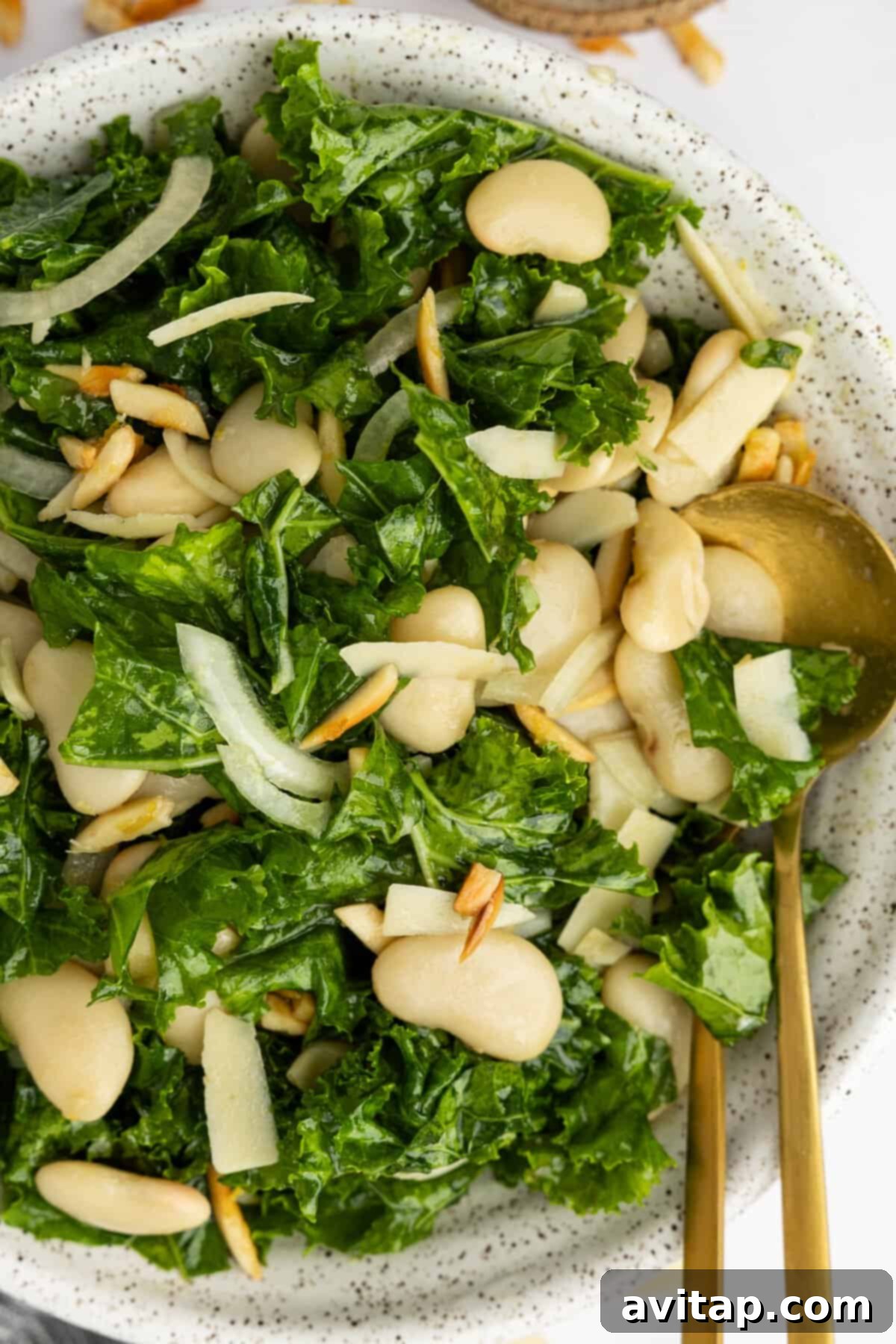 A bowl of kale salad with white beans, sliced onions, slivered almonds, and shaved cheese, served with a gold fork and spoon.