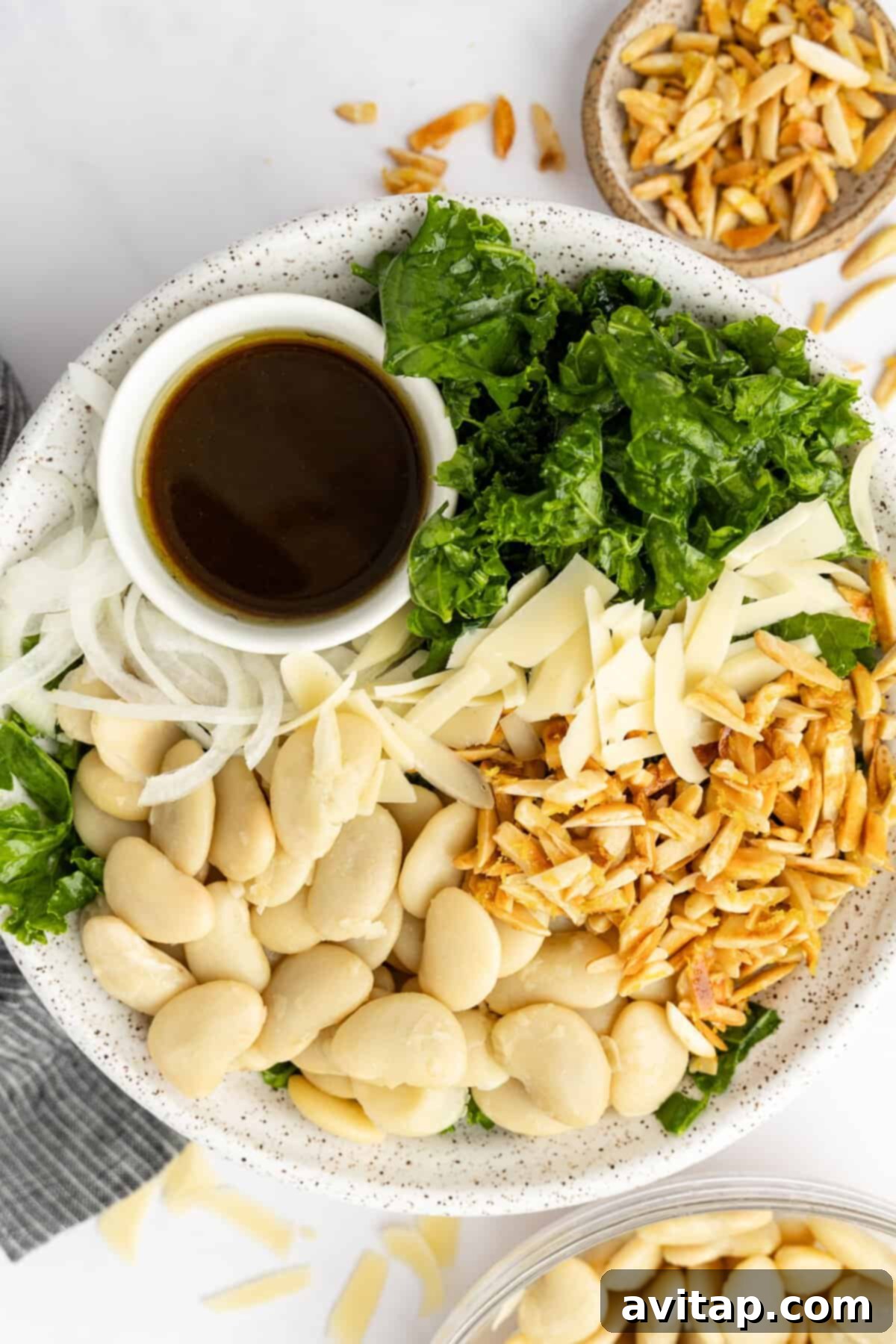 A bowl containing kale, sliced onions, butter beans, slivered almonds, shredded cheese, and a small dish of dark dressing.