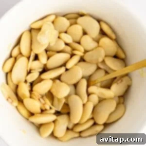 A white bowl filled with cooked butter beans and a gold spoon on a white surface.
