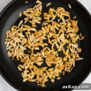 A black skillet with slivered almonds being toasted to a golden brown on a white surface.