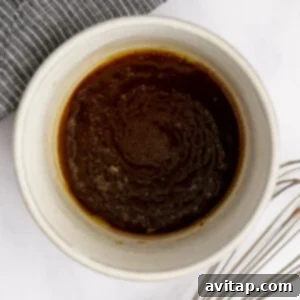 A white bowl containing a dark brown liquid mixture with a whisk placed nearby on a white surface, next to a gray-striped cloth.