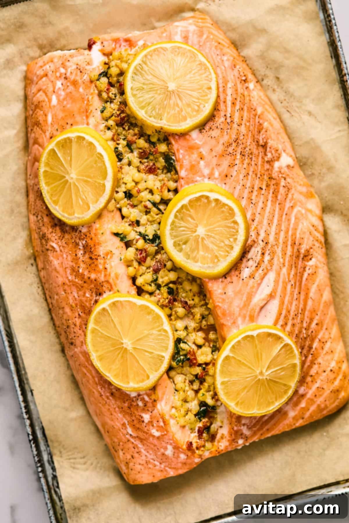 Baked salmon fillet stuffed with a vibrant pesto couscous mixture, elegantly topped with fresh lemon slices, resting on parchment paper in a baking tray.