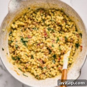 A vibrant bowl filled with a fresh couscous salad, richly mixed with herbs, spices, and chopped sun-dried tomatoes, with a wooden spoon resting inside, all on a marble surface.