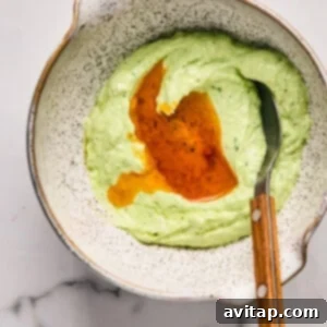 A small bowl of vibrant green pesto being thinned with a drizzle of sun-dried tomato oil, stirred with a spoon, on a textured background.
