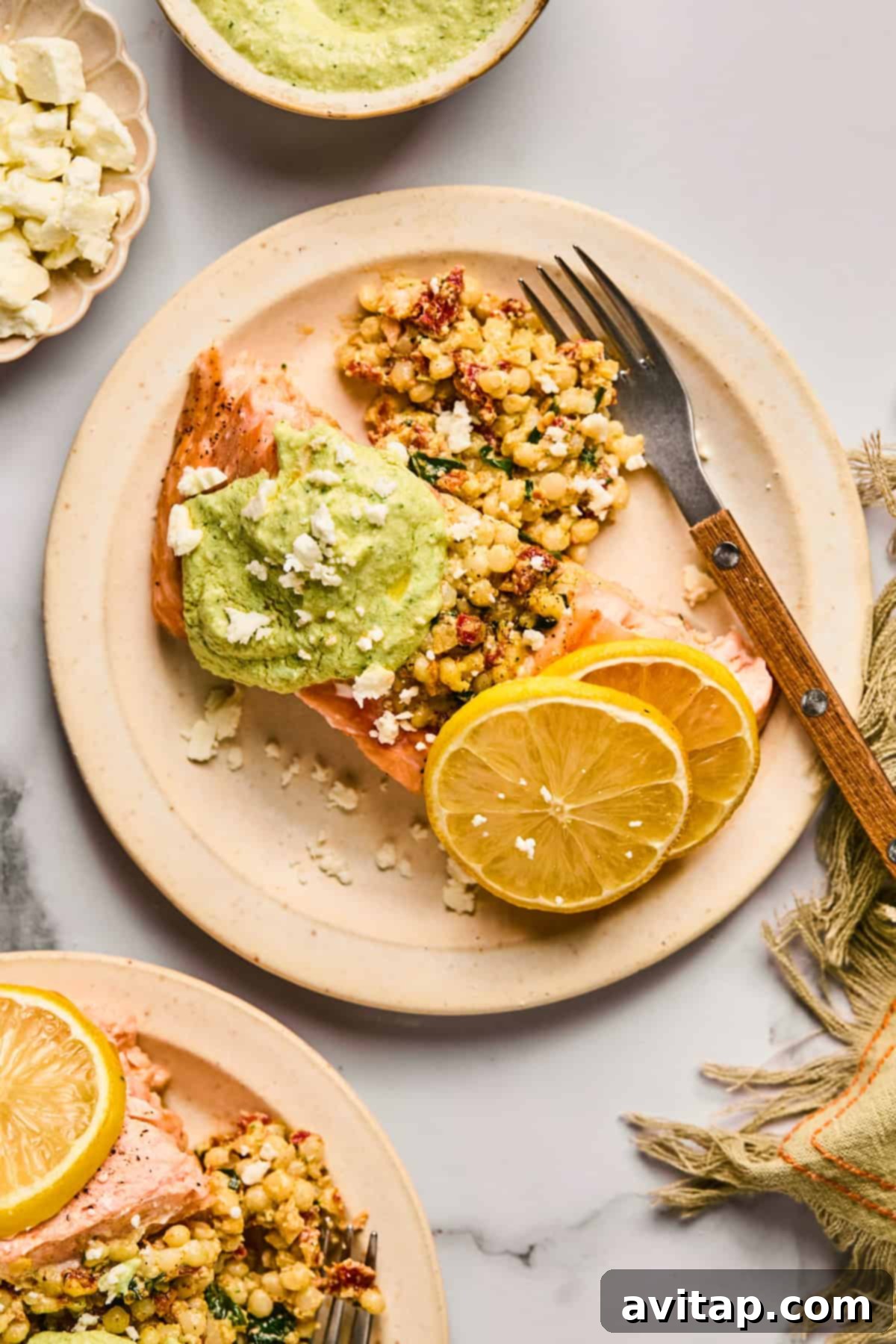 A stunning plate of baked salmon, generously topped with vibrant green pesto, accompanied by a side of fluffy couscous, crumbled feta cheese, and fresh lemon slices. A fork rests on the plate, with a linen napkin beside it, suggesting a ready-to-eat meal.