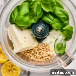 A food processor containing fresh basil leaves, crumbled cheese, pine nuts, and lemon halves, ready to be blended into a vibrant pesto on a marble surface.