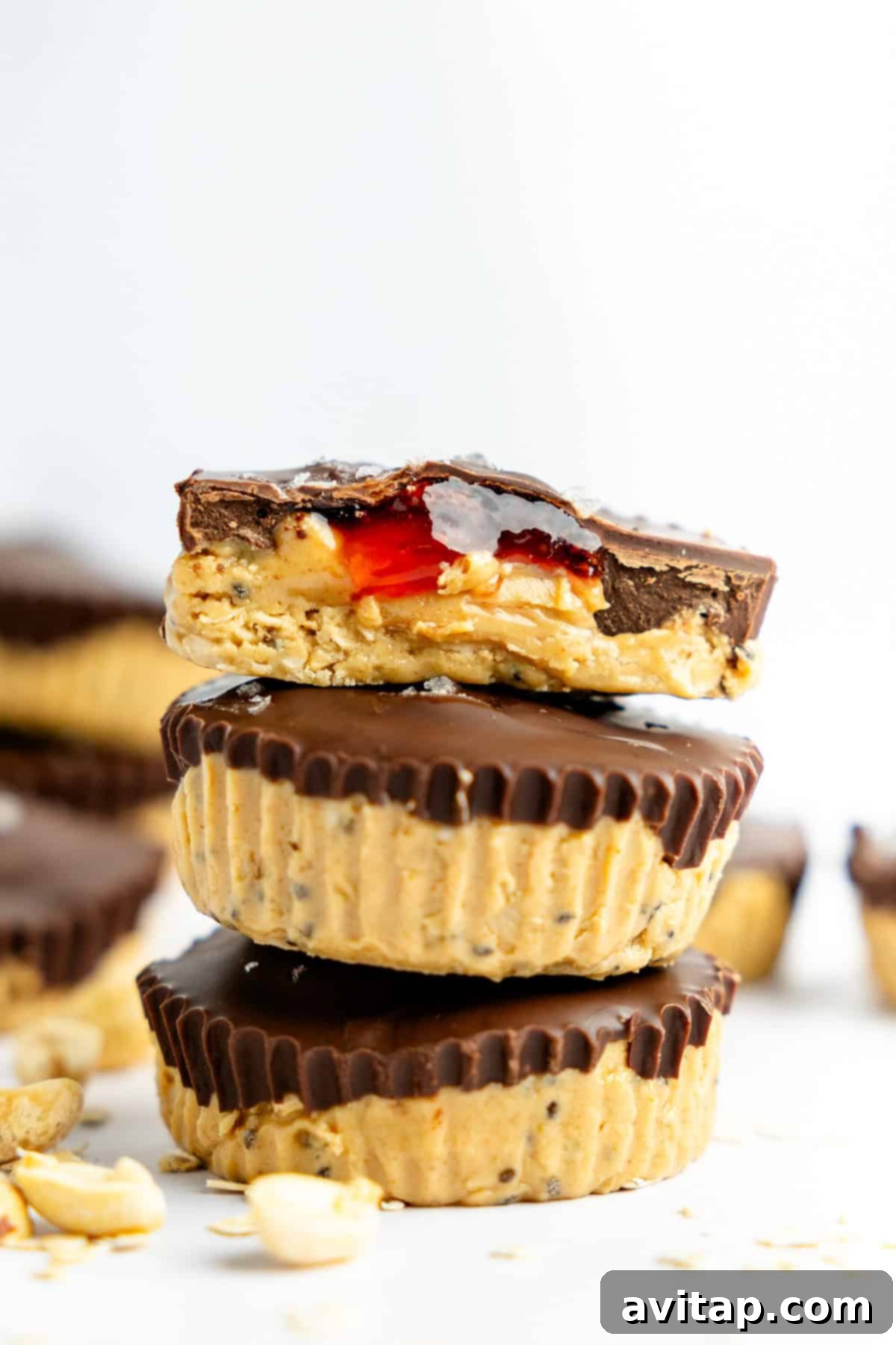 Three decadent peanut butter and chocolate cups stacked neatly, with the top cup revealing a vibrant red jam and creamy peanut butter filling after a bite has been taken. Loose peanuts are scattered around the base, hinting at the key ingredient.