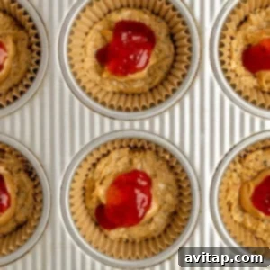 Muffin tin with paper liners, each containing an oat base, a layer of peanut butter, and a bright red dollop of jam, poised for the final topping.