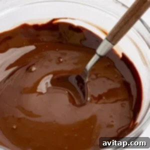 A clear glass bowl filled with rich, glossy melted chocolate, featuring a silver spoon resting inside, ready to be used for topping.