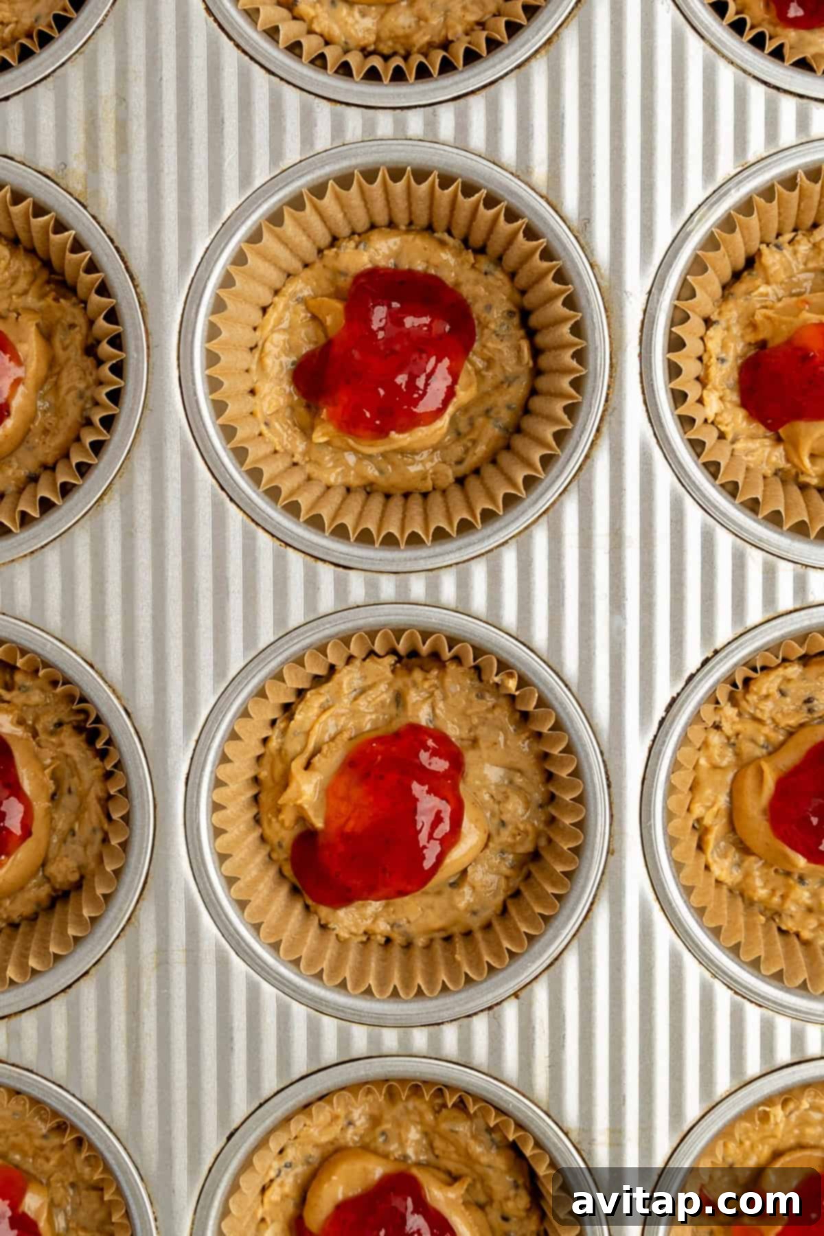 A muffin tin lined with paper baking cups, each filled with a layered base of unbaked oat mixture, topped with a dollop of creamy peanut butter, and finished with a spoonful of vibrant red jam, ready to be chilled.