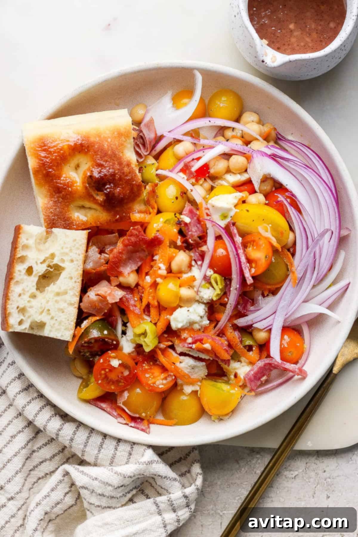 A vibrant no-lettuce Italian chopped salad featuring cherry tomatoes, red onion, shredded carrots, crispy bacon, crumbled cheese, and chickpeas, served alongside two pieces of crusty bread. A small cup of zesty dressing is nearby.
