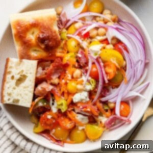A beautifully presented salad featuring cherry tomatoes, red onion, shredded carrots, crispy bacon bits, crumbled cheese, and chickpeas, elegantly arranged in a bowl. Accompanying the salad are two slices of rustic bread and a small condiment cup filled with dressing.