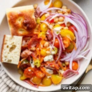 A finished bowl of No Lettuce Italian Chopped Salad featuring cherry tomatoes, red onion, shredded carrots, crispy bacon-like prosciutto, mozzarella cheese, and garbanzo beans. Two pieces of bread are presented alongside, with a small cup of dressing.