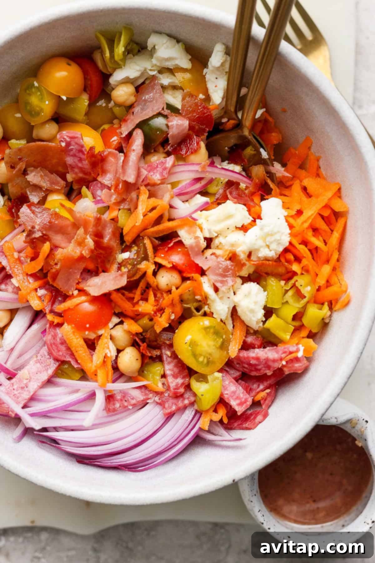 A close-up of a colorful No-Lettuce Italian Chopped Salad in a bowl, featuring perfectly sliced red onions, juicy cherry tomatoes, hearty chickpeas, finely shredded carrots, crumbled cheese, and generous pieces of cured meats. Two forks are elegantly placed in the bowl, ready for serving. A small, inviting bowl of dressing sits beside it.
