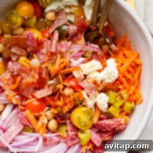 A vibrant salad bowl with shredded carrots, sliced red onions, cherry tomatoes, chickpeas, crumbled feta cheese, chopped bacon, and salami. Two forks are placed in the bowl, inviting a taste.