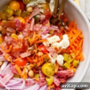 A tantalizing salad bowl featuring shredded carrots, thinly sliced red onions, juicy cherry tomatoes, hearty chickpeas, crumbled feta cheese, crispy chopped bacon, and flavorful salami pieces. Two forks are invitingly placed in the bowl.