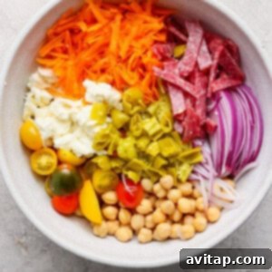 A bowl brimming with colorful ingredients for the No-Lettuce Italian Chopped Salad, including shredded carrots, sliced salami, red onions, chickpeas, cherry tomatoes, mozzarella cheese, and vibrant chopped green peppers, all ready to be mixed.