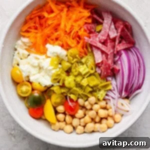 A large mixing bowl filled with the colorful salad base ingredients: shredded carrots, sliced salami, red onions, chickpeas, vibrant cherry tomatoes, creamy mozzarella, and chopped green peppers.
