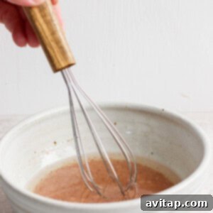 A hand expertly using a wooden-handled whisk to stir and combine liquid ingredients in a rustic ceramic bowl, creating the flavorful dressing for the salad.