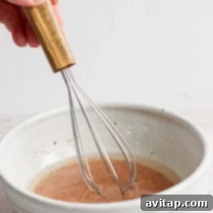 A hand holds a wooden-handled whisk, stirring the liquid ingredients vigorously in a ceramic bowl to create the zesty dressing.