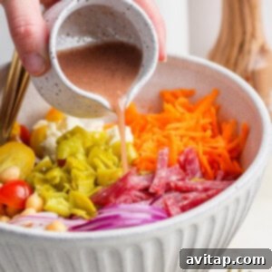 A hand gently pours a rich dressing onto a colorful salad, which includes bright red tomatoes, chickpeas, savory salami, thinly sliced red onions, tangy pepperoncini, shredded carrots, and crumbled feta cheese, all nestled in a large bowl. A serving fork and a pepper grinder are visible nearby.