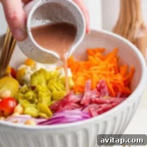 A hand carefully pours the zesty dressing onto the vibrant salad, which includes tomatoes, chickpeas, salami, red onions, pepperoncini, shredded carrots, and feta cheese in a large bowl. A fork and pepper grinder are poised nearby.