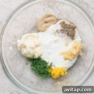 Glass bowl with Greek yogurt, mayo, Dijon mustard, minced garlic, chopped dill, lemon zest, black pepper, and salt on a textured surface.