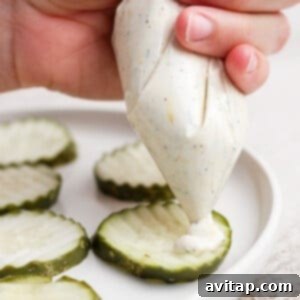 A hand squeezes creamy sauce from a piping bag onto crinkle-cut pickle slices arranged on a white plate.