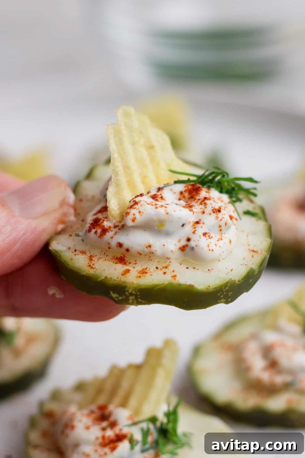 A hand holding a sliced cucumber topped with a dollop of cream, a sprinkle of paprika, a piece of ridged potato chip, and a sprig of dill.