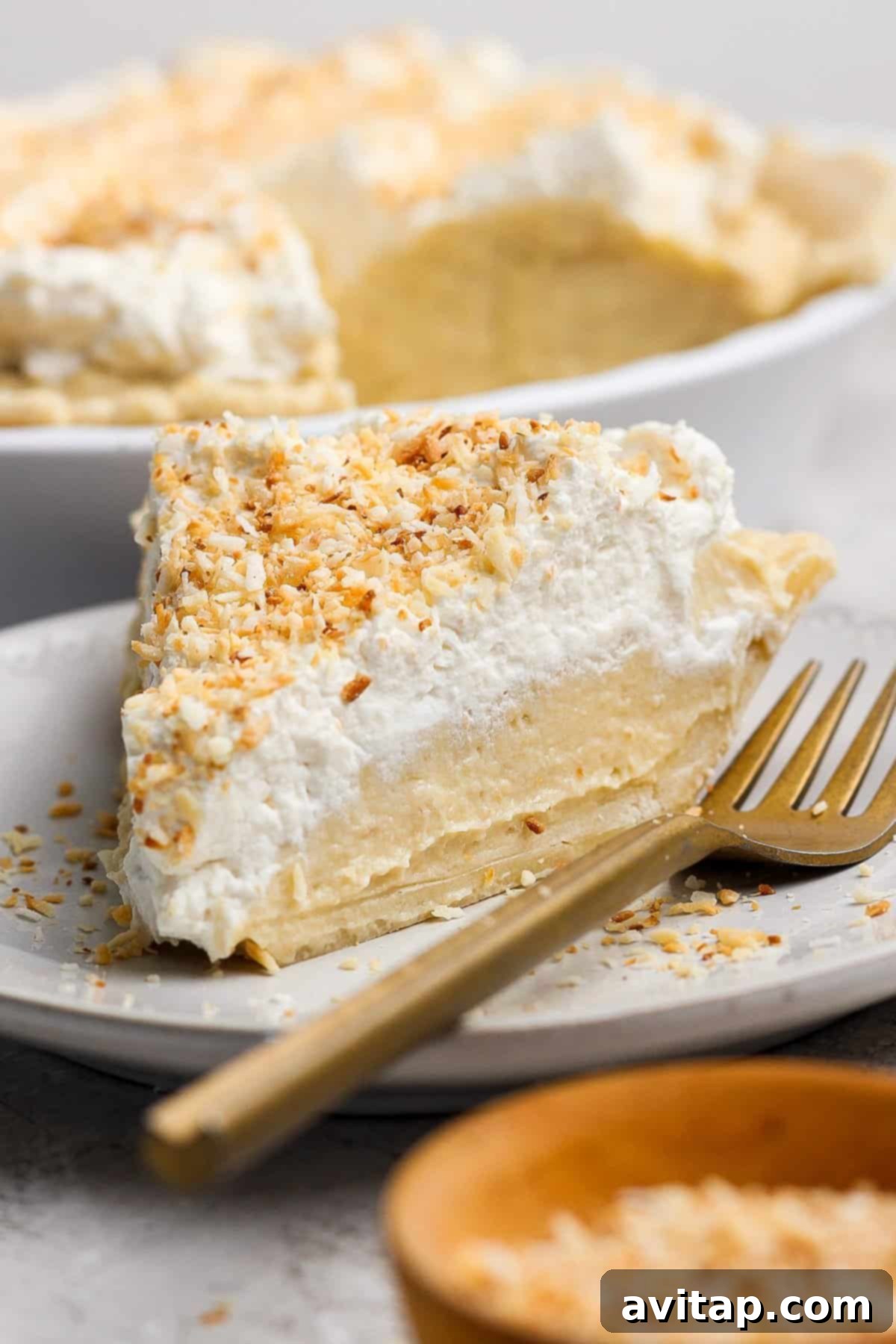 A beautifully sliced coconut cream pie, garnished with fluffy whipped cream and golden toasted coconut flakes, presented on a white plate with an elegant gold fork.