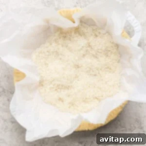 A round baking dish, meticulously lined with parchment paper and filled with uncooked white rice, rests on a subtly textured surface, demonstrating a method for blind baking.