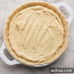 An unbaked pie crust, delicately filled with a pale, creamy batter, rests invitingly in a pristine white baking dish, set upon a textured surface, ready for the oven.