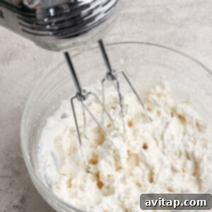 An electric mixer actively whipping cream in a clear glass bowl, positioned on a light-colored kitchen counter, creating fluffy peaks.