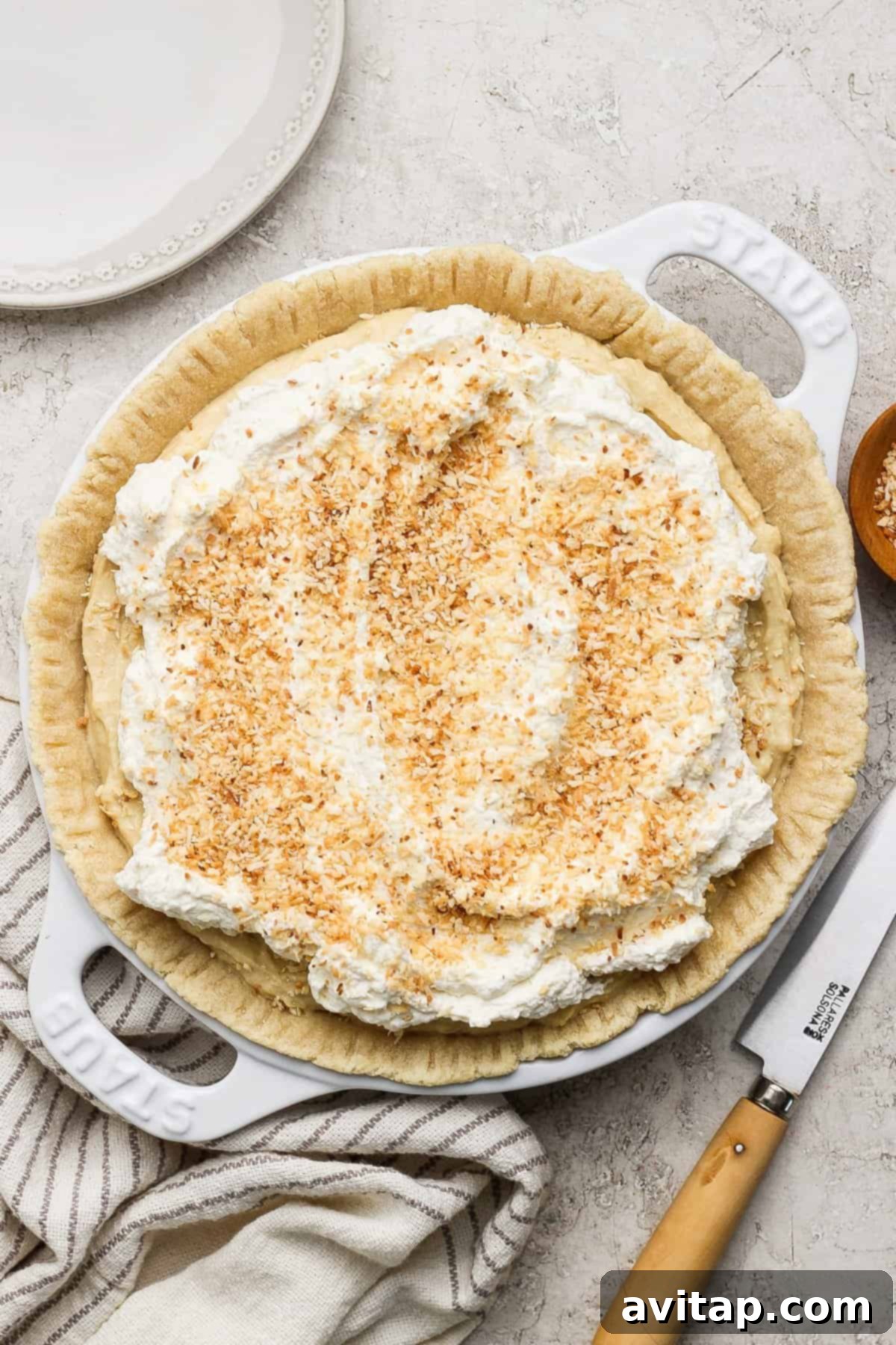 A perfectly baked pie, generously topped with swirls of whipped cream and a sprinkle of golden toasted coconut flakes, presented on a light-colored surface beside a polished serving knife and a neatly folded striped cloth.