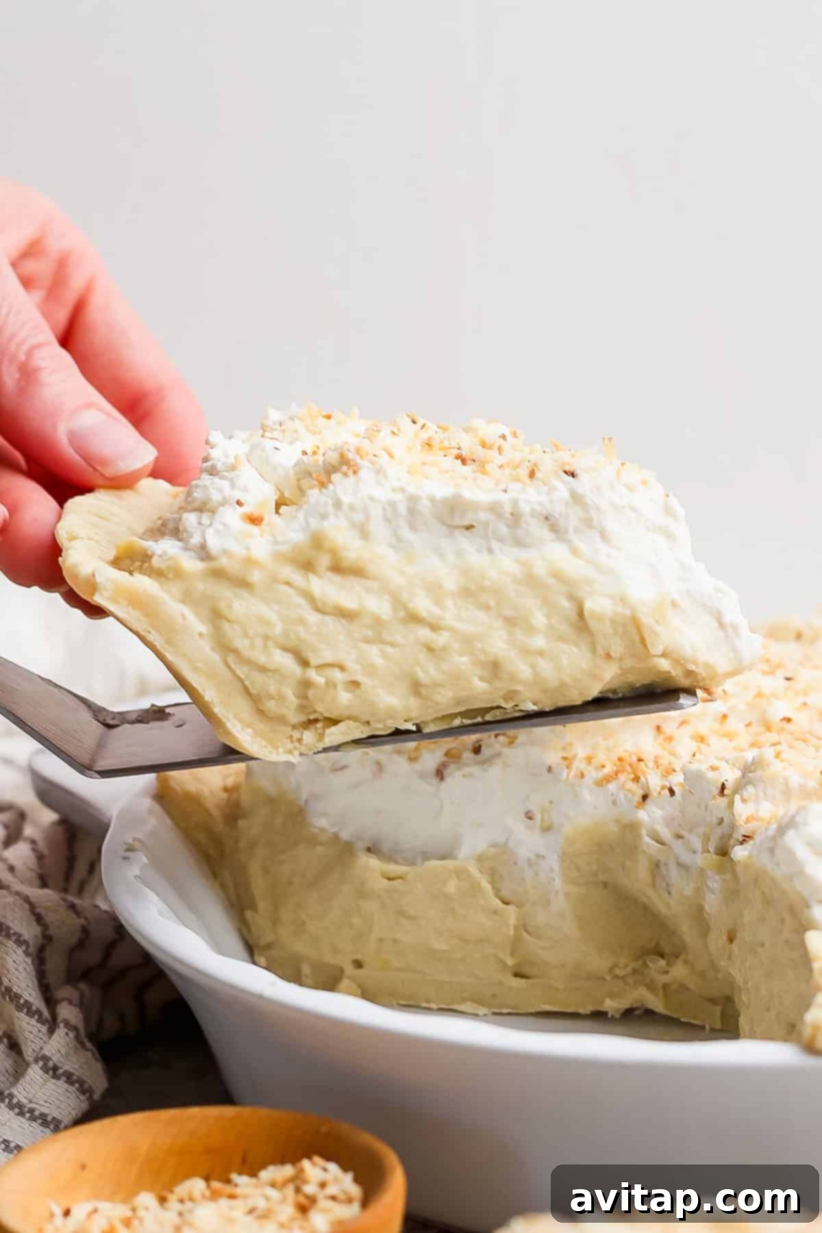 A hand gracefully serves a generous slice of creamy pie, adorned with a dollop of whipped cream and a delicate sprinkling of toasted coconut, from a pristine white pie dish.