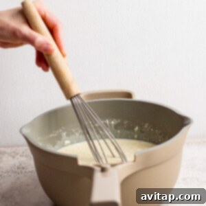 A person expertly whisks a creamy batter within a beige mixing bowl, set against a pristine light-colored countertop, capturing the motion and texture of the preparation.