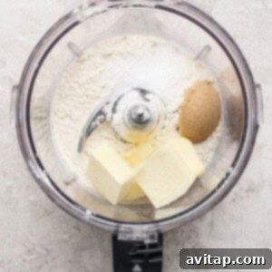 The bowl of a food processor containing a blend of white flour, golden brown sugar, sea salt, and cold, cubed butter, ready for processing, positioned on a light countertop.