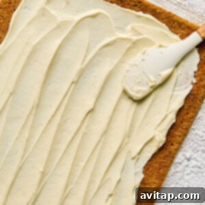 A rectangular cake layer being spread with cream cheese frosting using a spatula on a floured surface.
