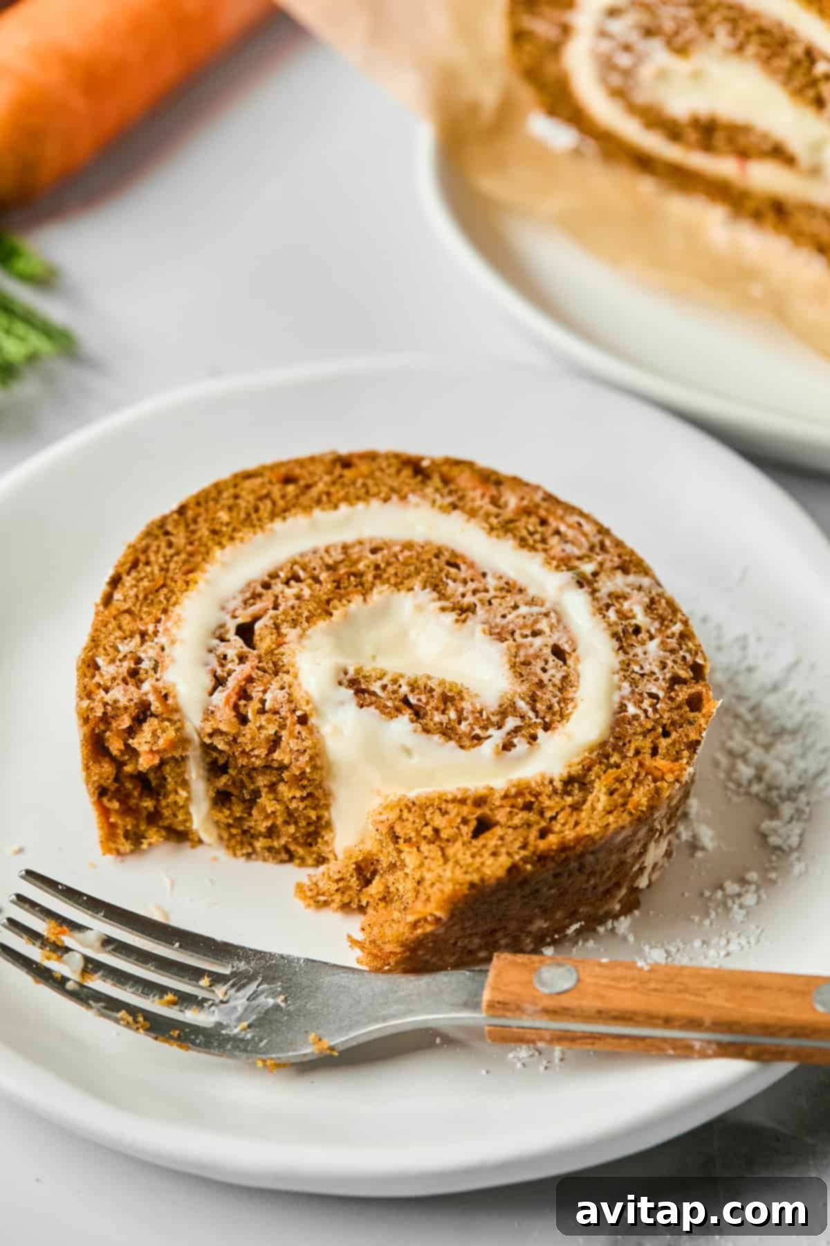 A perfectly sliced piece of protein carrot cake roll on a white plate, revealing layers of moist cake and creamy filling, with a fork nearby.