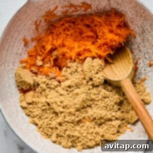 A mixing bowl containing shredded carrots and brown sugar, with a wooden spoon resting inside.