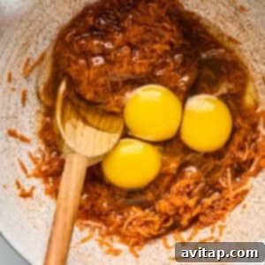 Three raw eggs are cracked into a mixing bowl containing brown shredded ingredients, with a wooden spoon partially submerged in the mixture.