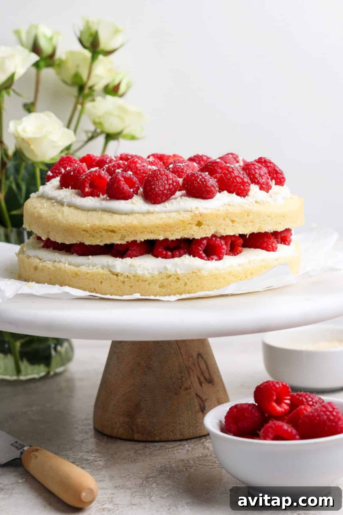 Two-layer coconut raspberry cake with white frosting and fresh raspberries on top, presented beautifully on a wooden cake stand. The background features delicate white flowers and a bowl brimming with fresh raspberries, enhancing the visual appeal.