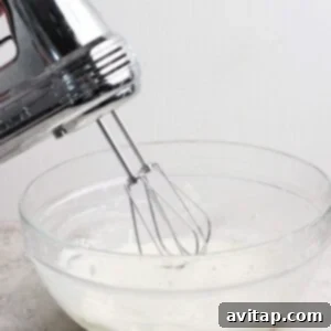 Electric hand mixer with beaters above a glass bowl of white liquid, possibly cream or batter, on a countertop.