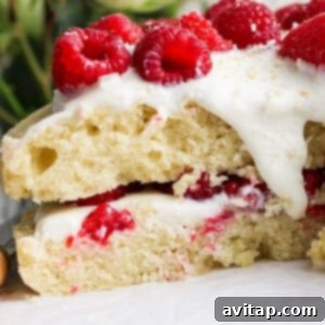 Close-up of a cake layered with white frosting and fresh raspberries, with a few white roses in the blurred background.