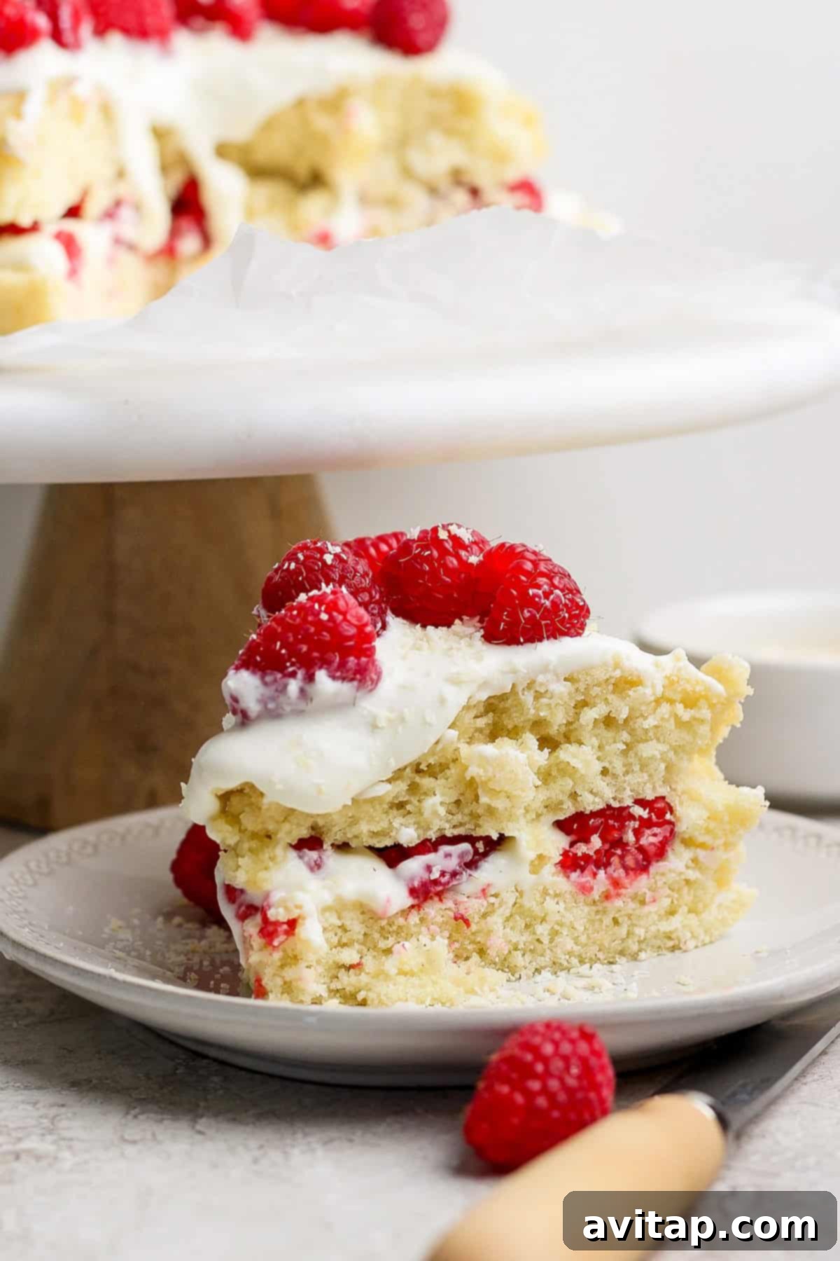 A perfectly sliced piece of vanilla coconut cake, revealing layers of white frosting and fresh raspberries within. It's elegantly presented on a plate with a wooden-handled knife resting beside it, inviting indulgence.