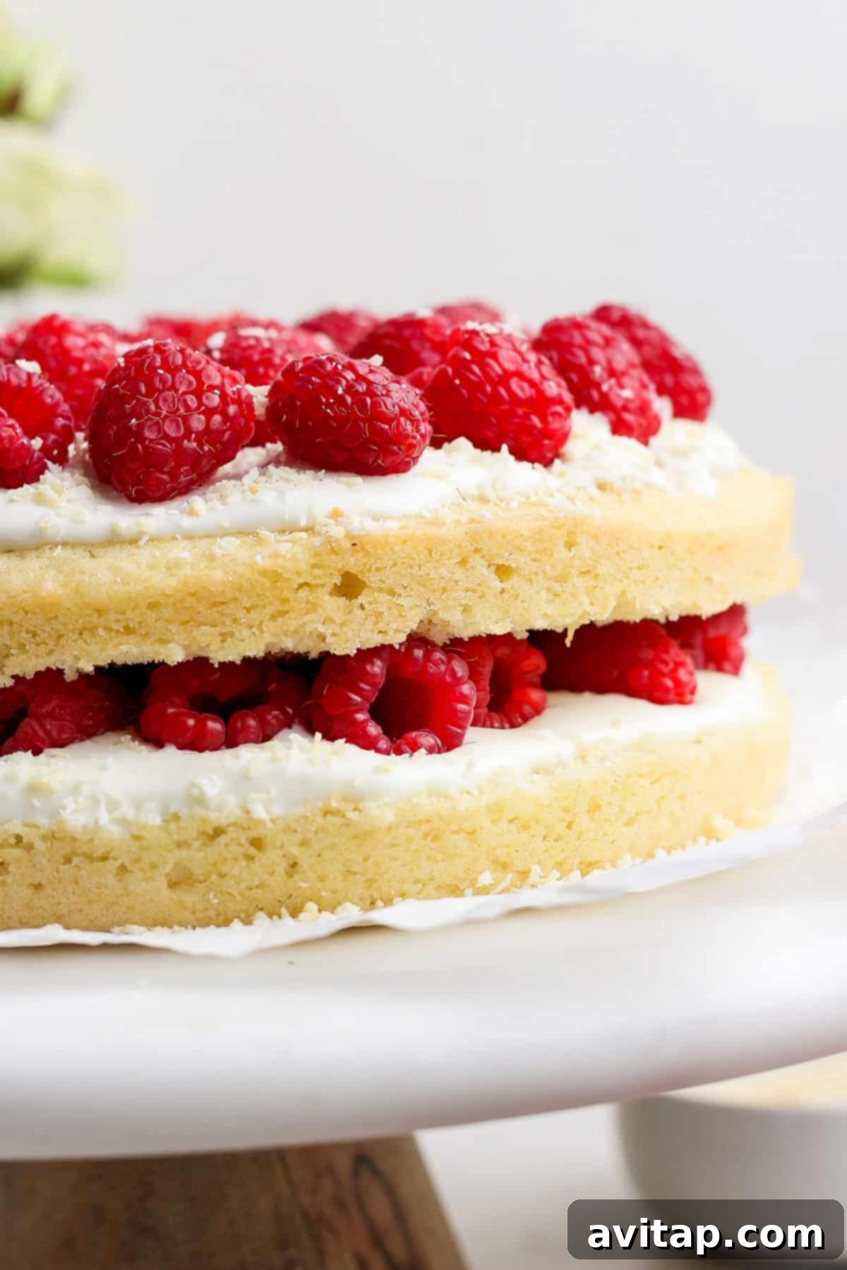 A stunning layered coconut cake, generously adorned with creamy white frosting and vibrant fresh raspberries, proudly displayed on a classic cake stand.