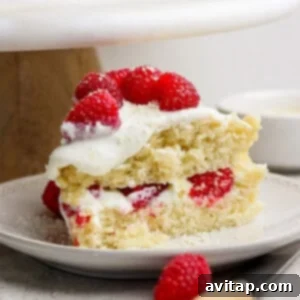 A slice of vanilla cake with cream and raspberries on a plate. A whole cake is partially visible in the background.