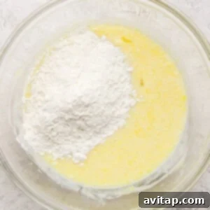 A glass bowl containing a mixture of yellow batter with a mound of white flour on top, set on a textured surface.