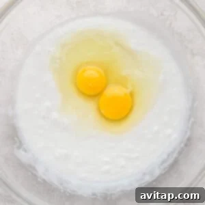 Two raw eggs cracked on top of a mixture in a clear glass bowl on a textured surface.